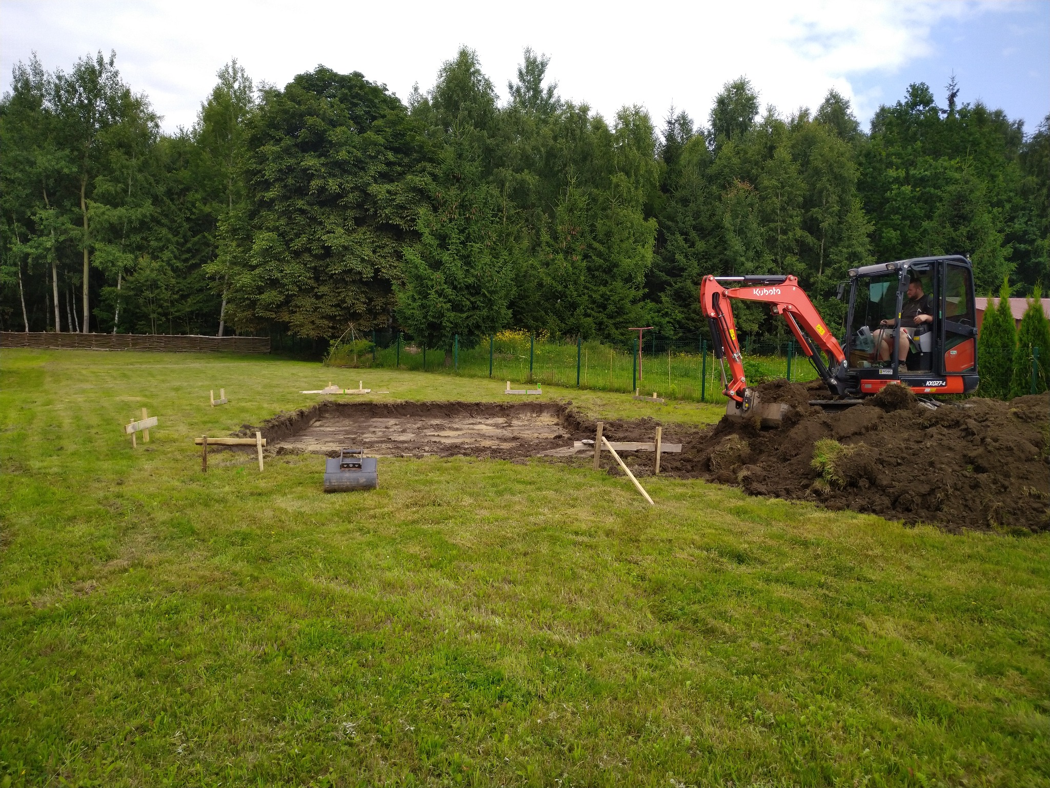 Pomarańczowa mini koparka Kubota w trakcie prac ziemnych, wykop pod przyszły budynek, widoczne drewniane znaczniki wyznaczające obrys fundamentów na trawiastym terenie, w tle zielony las.