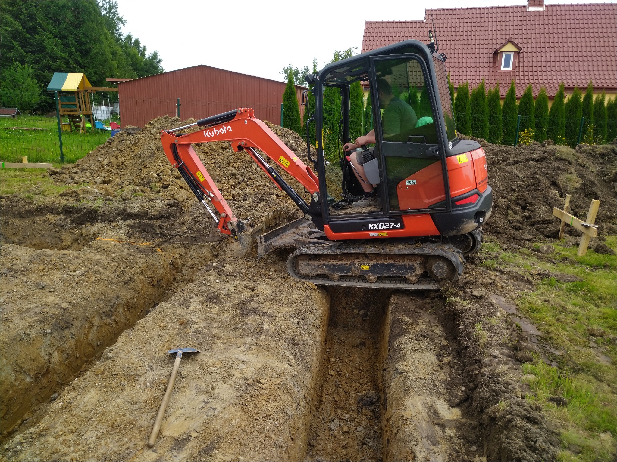 Pomarańczowa mini koparka Kubota KX027-4 w trakcie kopania wąskiego wykopu w ziemi, obok leży kilof, w tle dom z czerwoną dachówką i zielone tuje.