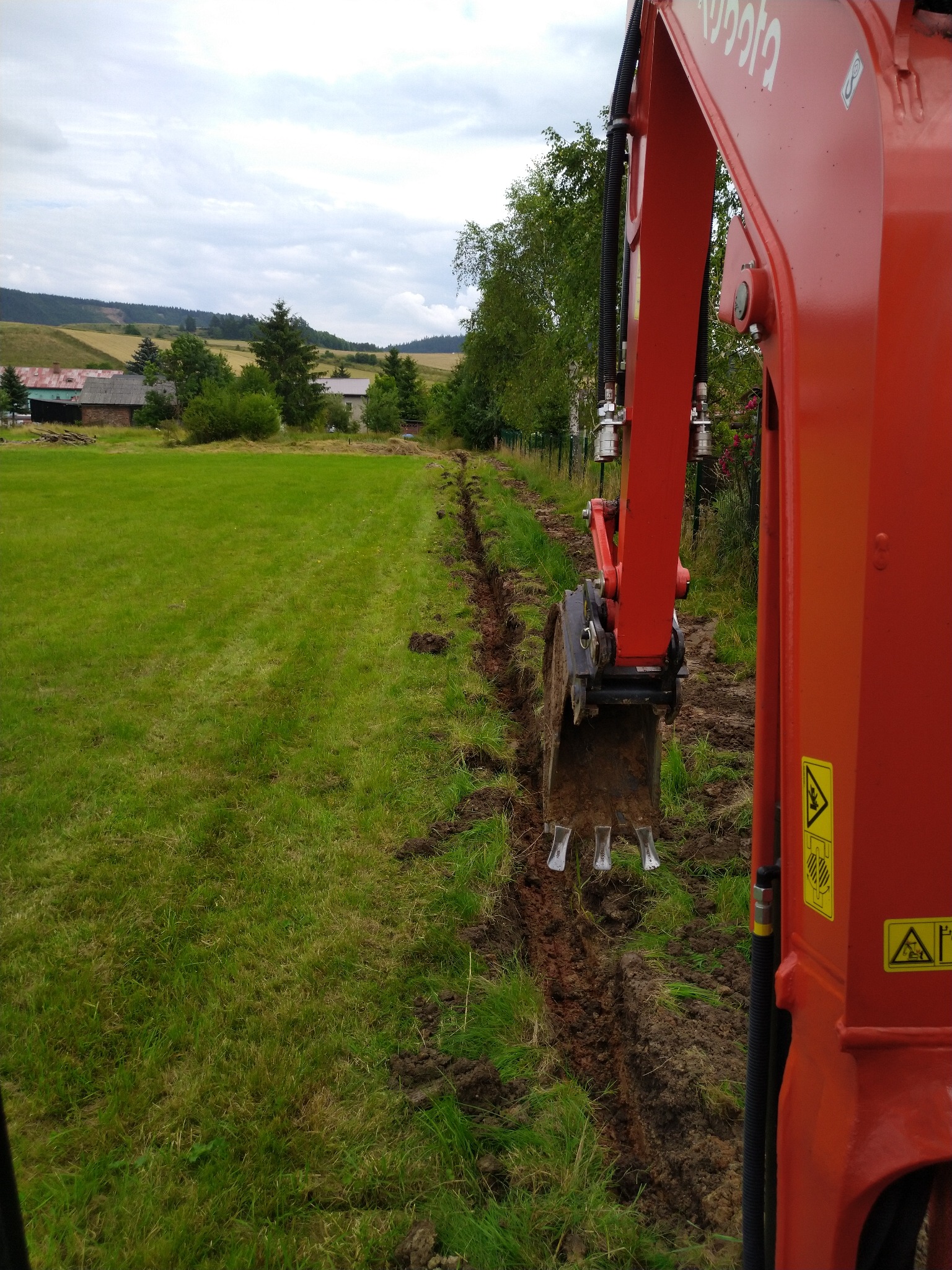 Pomarańczowa koparka Kubota częściowo widoczna, wykonująca wąski wykop na trawiastym terenie, w tle zabudowania wiejskie i wzgórza.