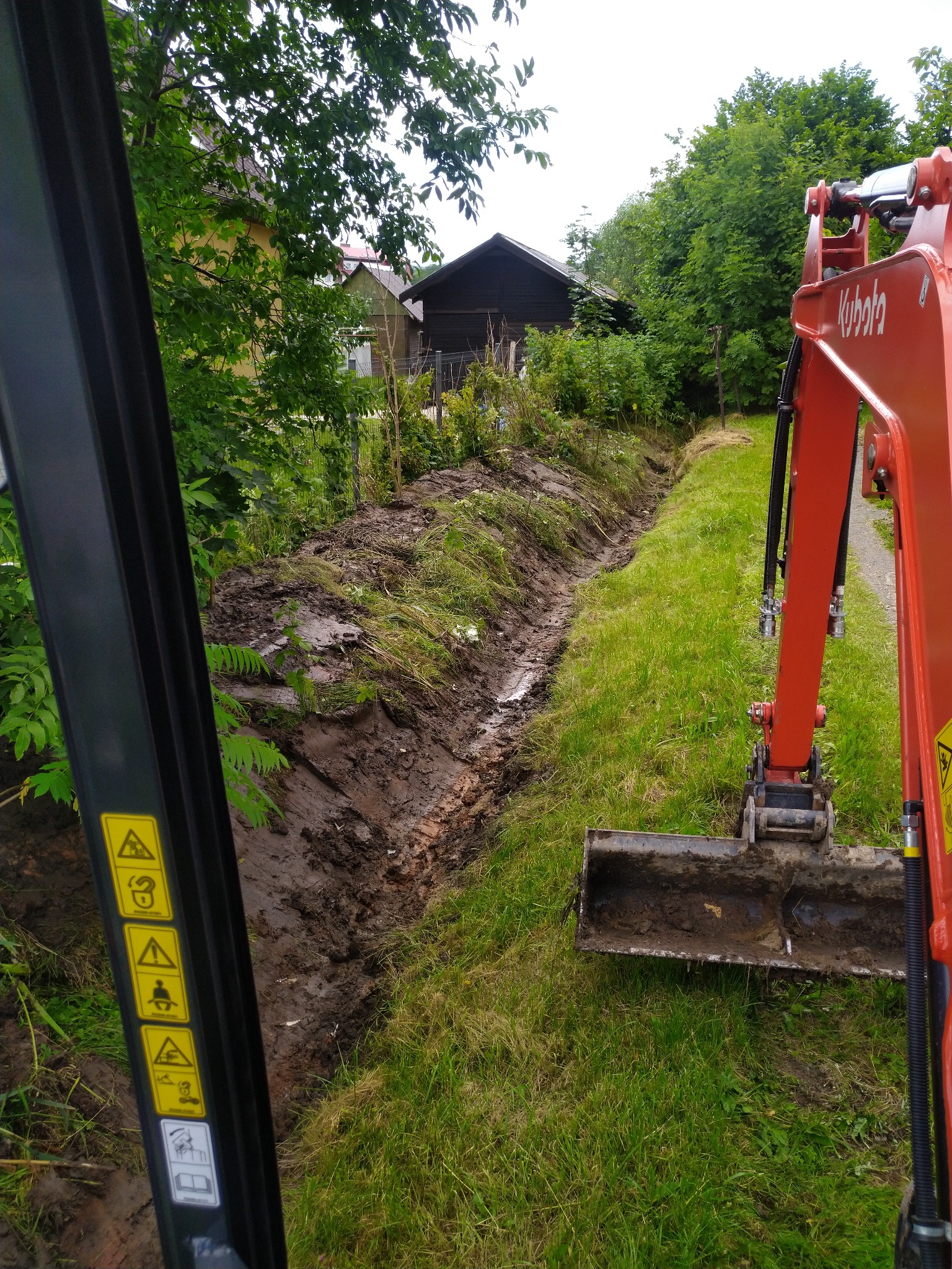 Wąski wykop w ziemi widziany z kabiny pomarańczowej mini-koparki Kubota, z widocznymi naklejkami ostrzegawczymi na czarnej ramie, trawa po bokach, w tle zabudowania.