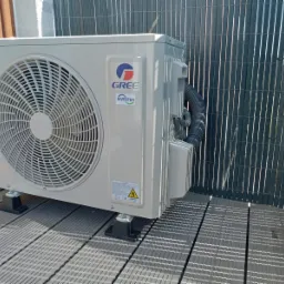 Zewnętrzna jednostka klimatyzatora GREE Inverter zamontowana na podwyższeniu z kompozytowych desek tarasowych, widoczna obok ażurowego ogrodzenia.