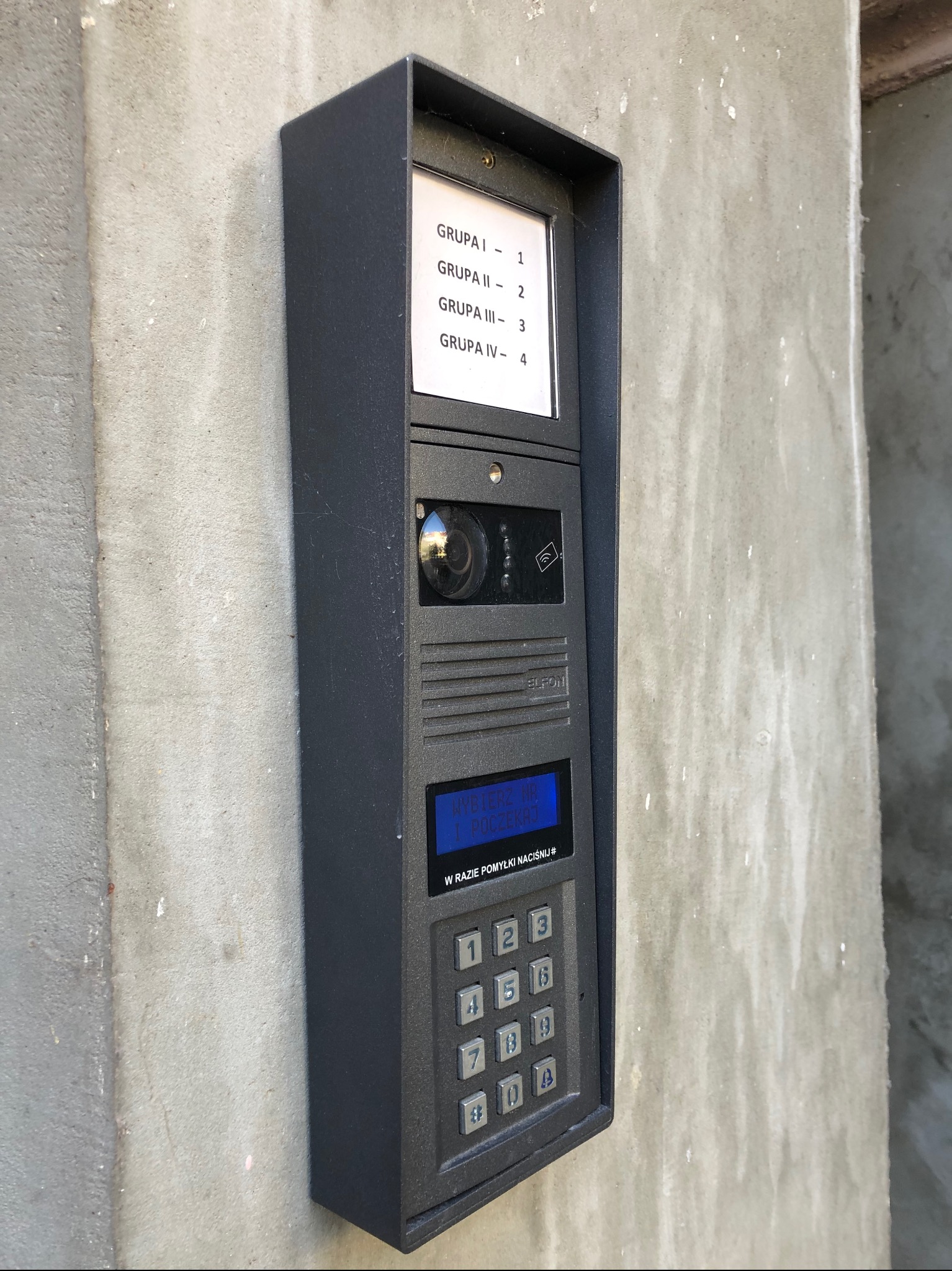 Czarny panel domofonowy z kamerą i klawiaturą numeryczną, zamontowany na szarej ścianie, z informacją o grupach lokatorów i instrukcją obsługi.