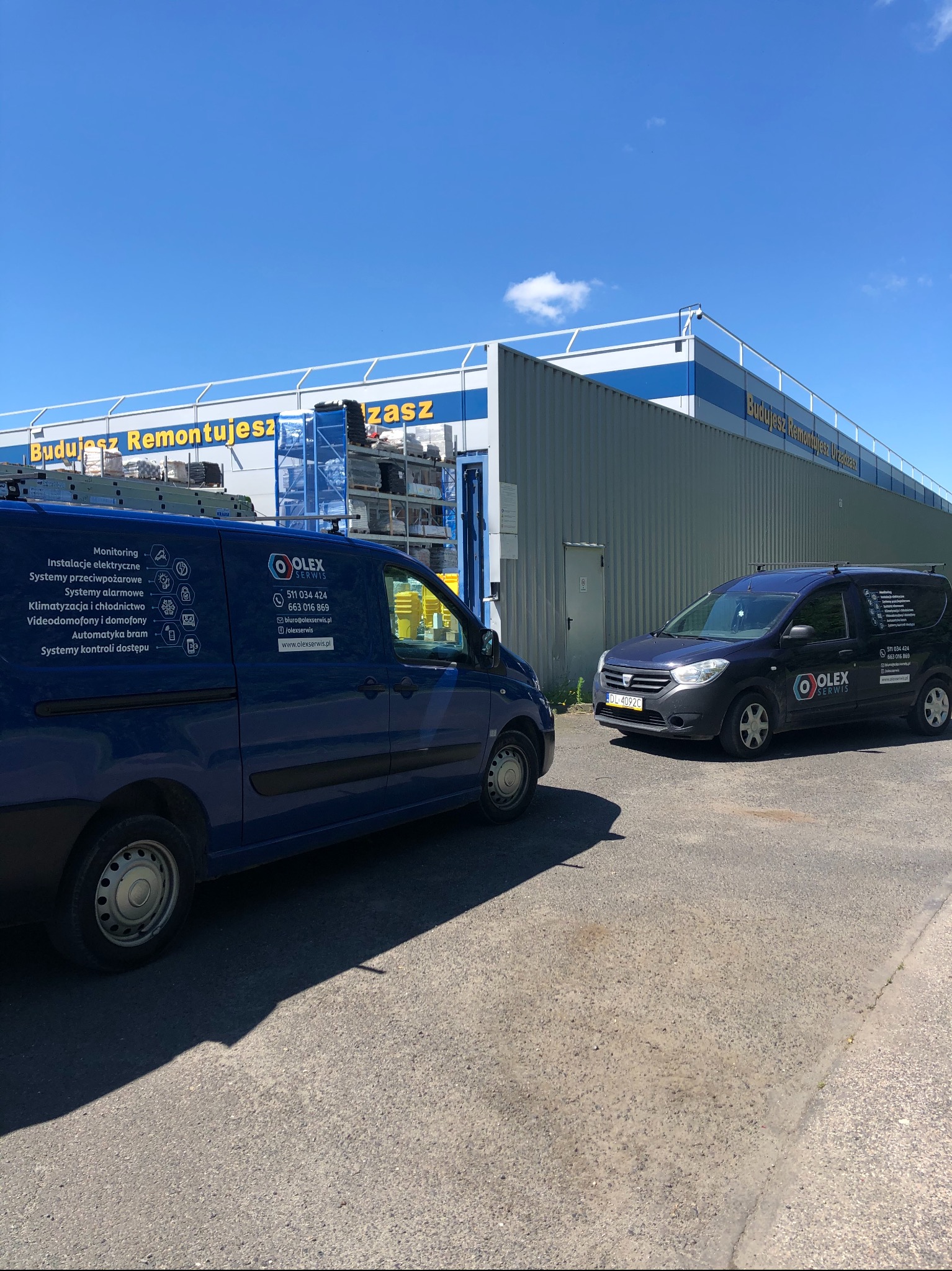 Dwa samochody dostawcze firmy Olex Serwis zaparkowane przed budynkiem z napisem 'Budujesz Remontujesz Urządzasz'. Na boku niebieskiego vana widoczne logo firmy i lista oferowanych usług: monitoring...