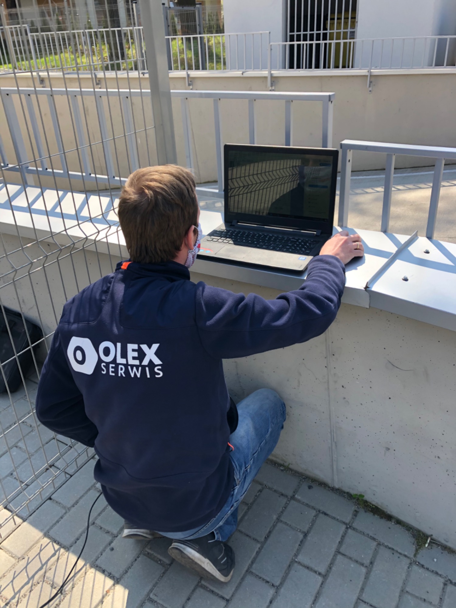 Instalator firmy Olex Serwis na zewnątrz budynku, konfigurujący system za pomocą laptopa na tle metalowej konstrukcji.