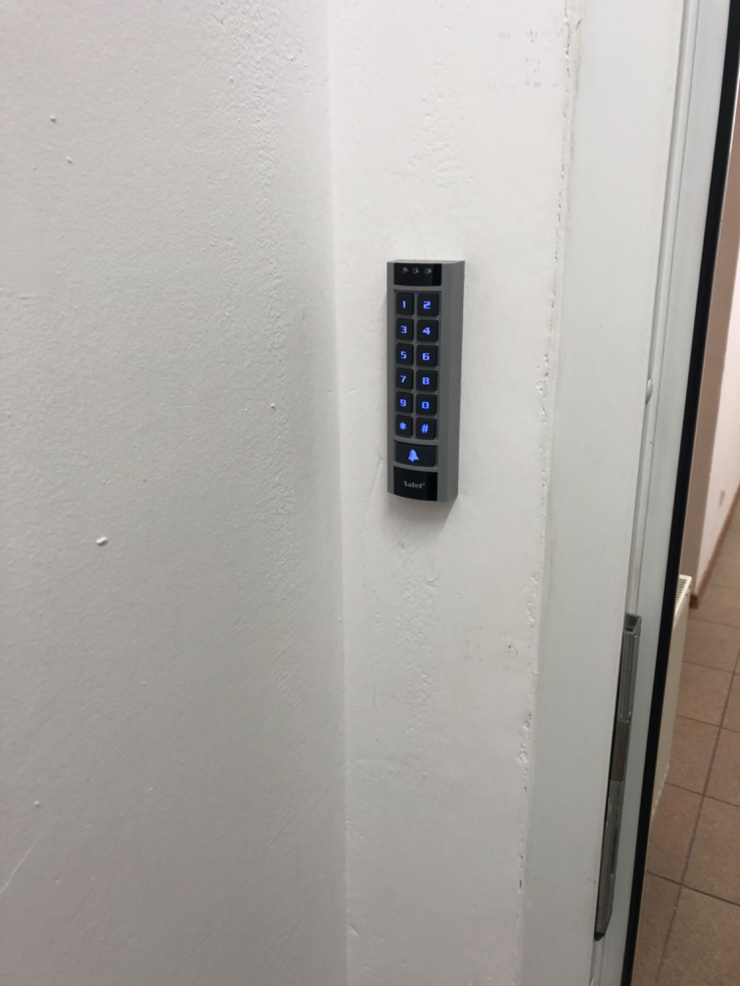 Szary panel domofonu cyfrowego z podświetlanymi na niebiesko przyciskami, zamontowany na białej ścianie przy futrynie drzwi.