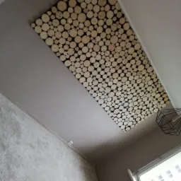 Dekoracyjny sufit z drewnianych krążków, widok z dołu, fragment lampy geometrycznej i okna w tle.