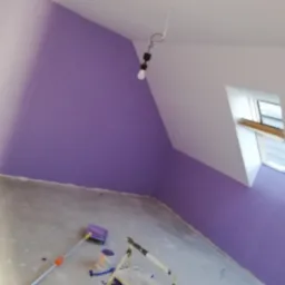 Ściana w pokoju na poddaszu pomalowana na fioletowo, widoczny wałek malarski, drabina i wiadro z farbą, okno dachowe w tle, surowy beton na podłodze.
