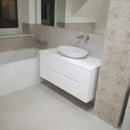 Biała, podwieszana szafka łazienkowa z umywalką nablatową, widoczna obok wanny i ozdobnej, beżowej płytki ściennej.