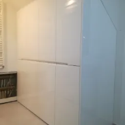 Duża, biała szafa z połyskiem, idealnie dopasowana do skosu dachu, z widocznym grzejnikiem i rurami w tle.