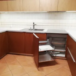 Kuchenna zabudowa w odcieniach brązu i beżu, z wysuwanymi koszami cargo w szafce narożnej, widok na płytki ścienne i blat zlewem.