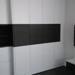 Biała szafa wnękowa z poziomym, czarnym pasem na frontach, dopasowana do ciemnej podłogi z desek.