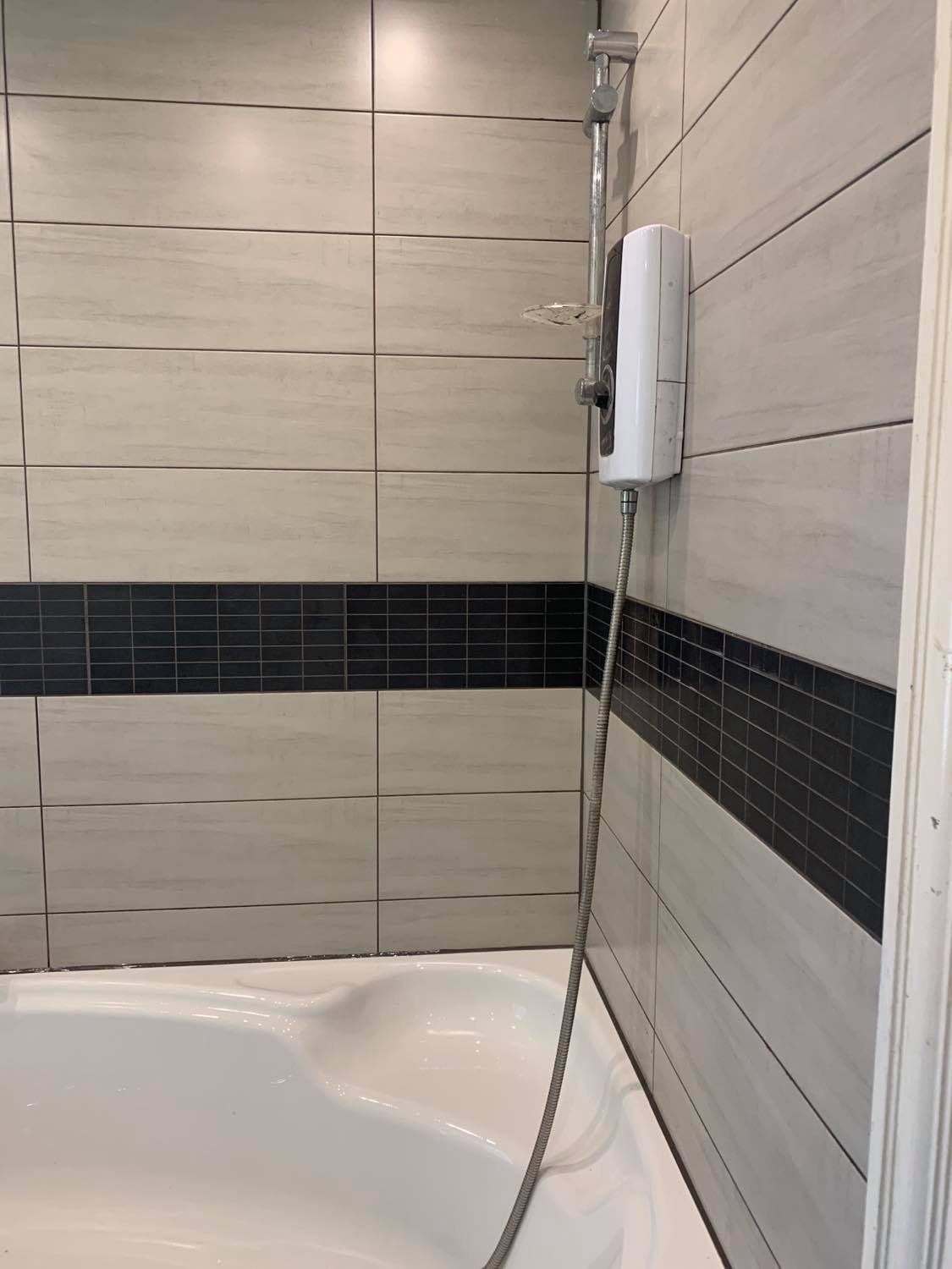 Biała wanna z zamontowanym prysznicem i podgrzewaczem wody na ścianie wyłożonej jasnymi płytkami z ciemnym pasem dekoracyjnym.