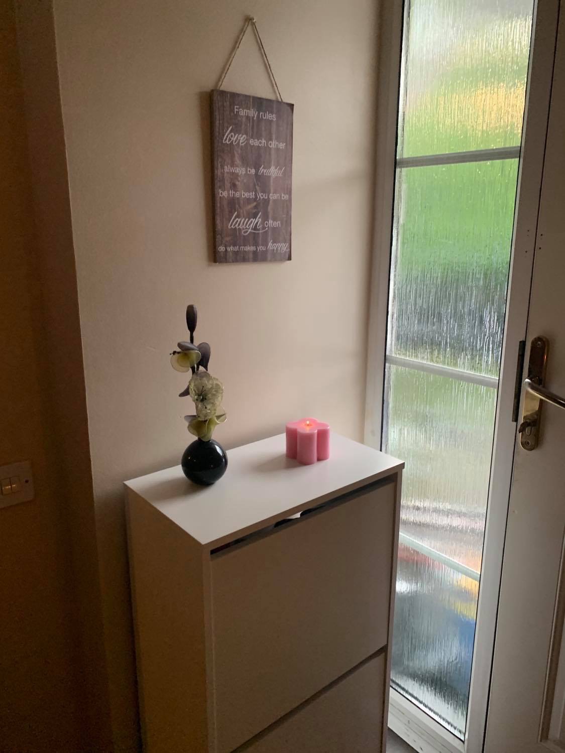 Przedpokój po remoncie: biała szafka na buty z dekoracjami (kwiaty w czarnym wazonie i różowa świeczka), na ścianie drewniana tabliczka z napisem 'Family rules', w tle przeszklone drzwi wejściowe.