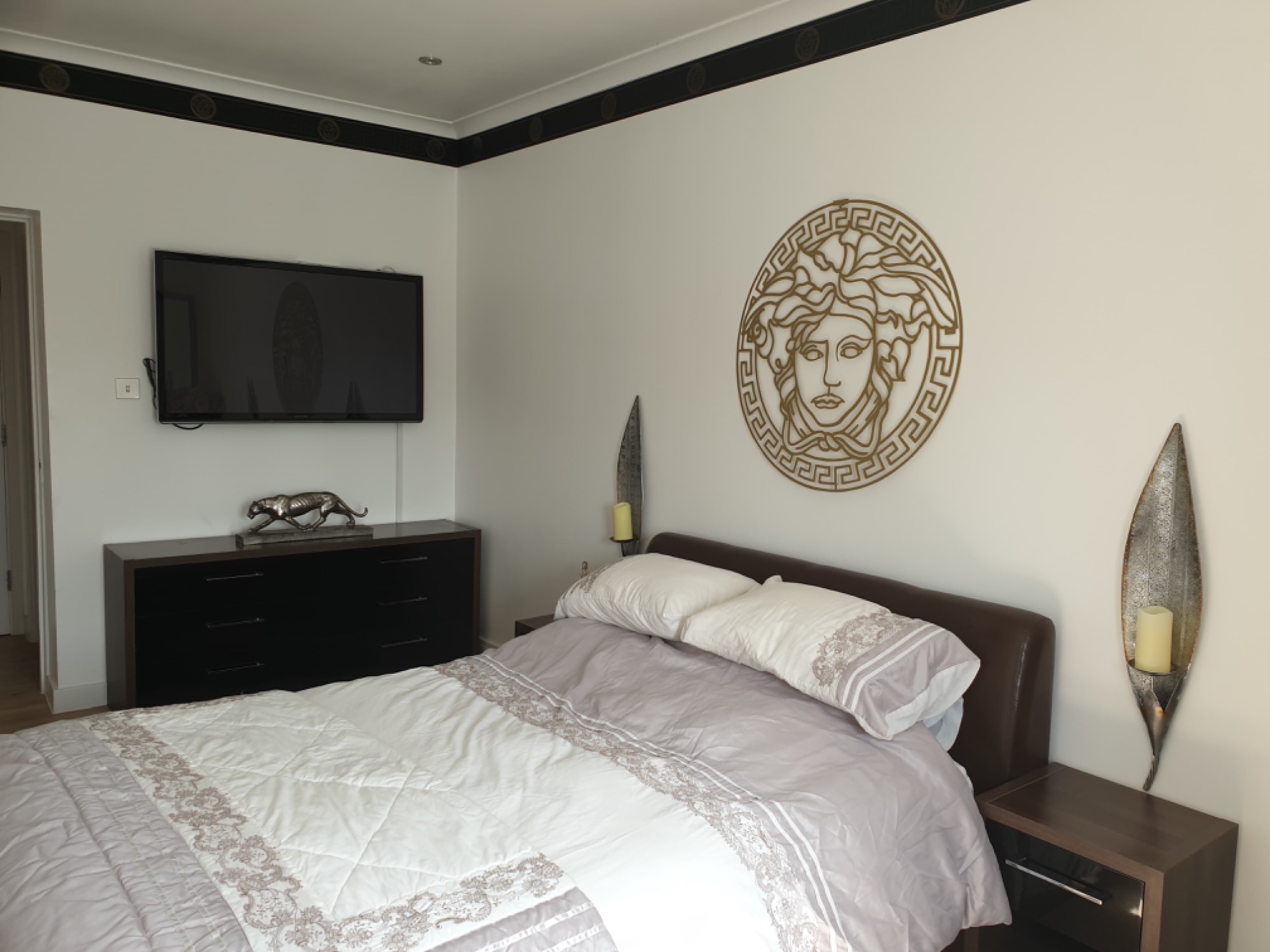 Sypialnia z łóżkiem, komodą z figurką pantery, telewizorem, dekoracją ścienną z motywem Meduzy i kinkietami w kształcie liści.