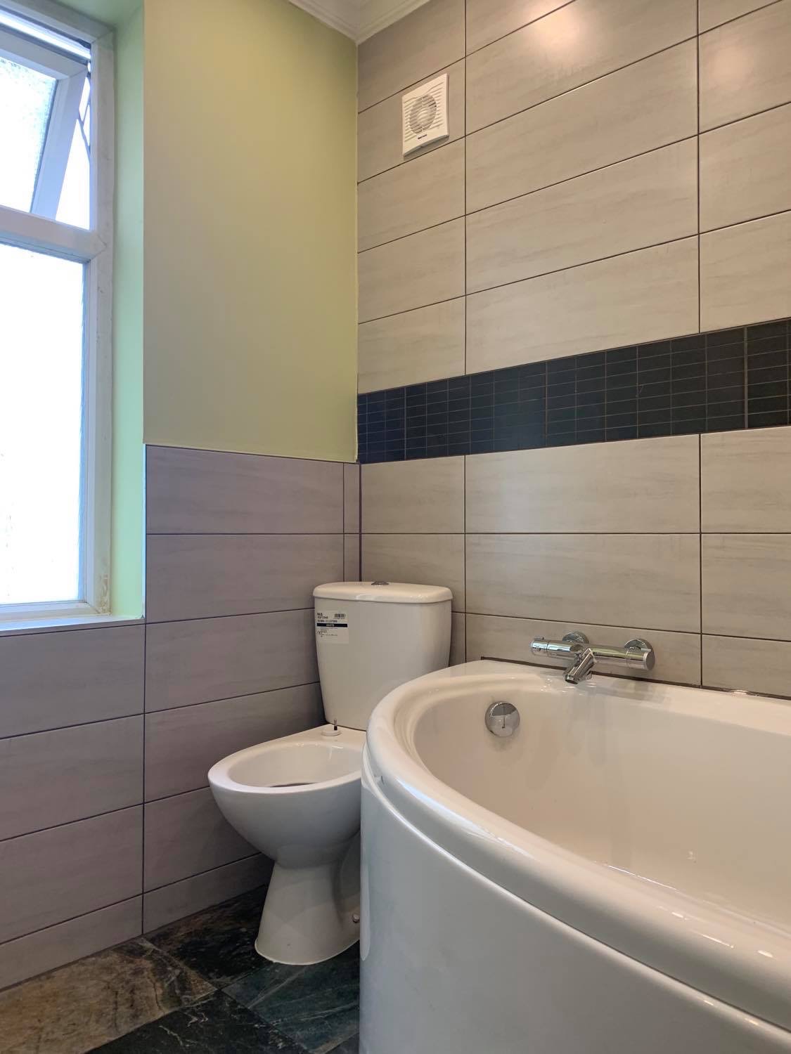 Wykończona łazienka z wanną narożną, toaletą i bidetem, wyłożona płytkami w odcieniach szarości z czarnym akcentem, widoczny wentylator na ścianie i okno z zielonym obramowaniem.