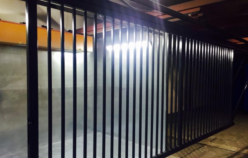 Klatka z pionowych, czarnych prętów, prawdopodobnie do zabezpieczenia przedmiotu podczas obróbki, widoczna żółta belka konstrukcyjna w tle.