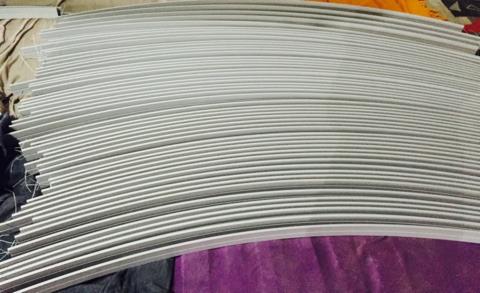 Stos wygiętych, szarych profili aluminiowych ułożonych warstwami na fioletowym i beżowym materiale.