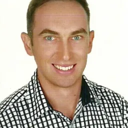 Portret uśmiechniętego mężczyzny w koszuli w kratę, z wyraźnymi zielonymi oczami, działającego jako specjalista w Garwolinie.