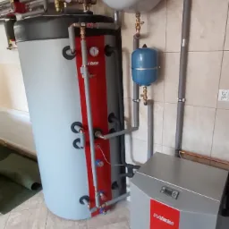 Szary zbiornik akumulacyjny ciepła z czerwonym pasem, obok szary kompaktowy moduł grzewczy z czerwonym pasem, rury instalacyjne, szara ściana z jasnymi płytkami w tle.