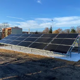 Rząd paneli słonecznych na metalowej konstrukcji, otoczony białym żwirem, na tle błękitnego nieba i zabudowań.