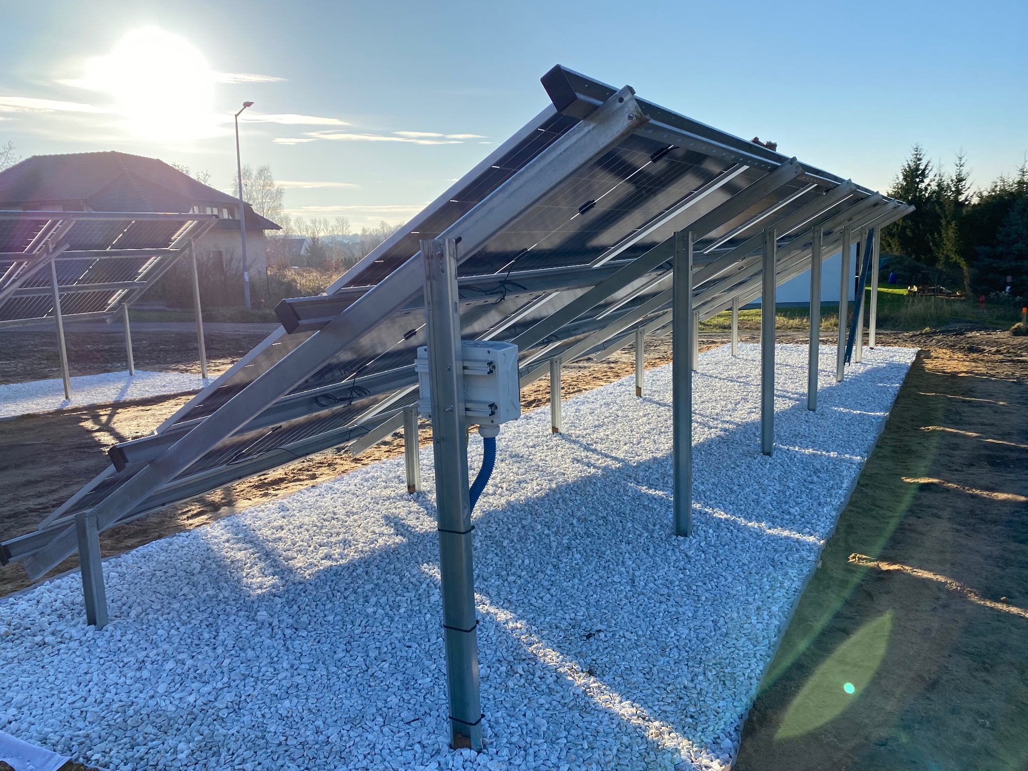 Montaż paneli słonecznych na konstrukcji wsporczej, widok od spodu, z widoczną instalacją elektryczną i białym żwirem wokół podstaw, w tle budynek mieszkalny i błękitne niebo.
