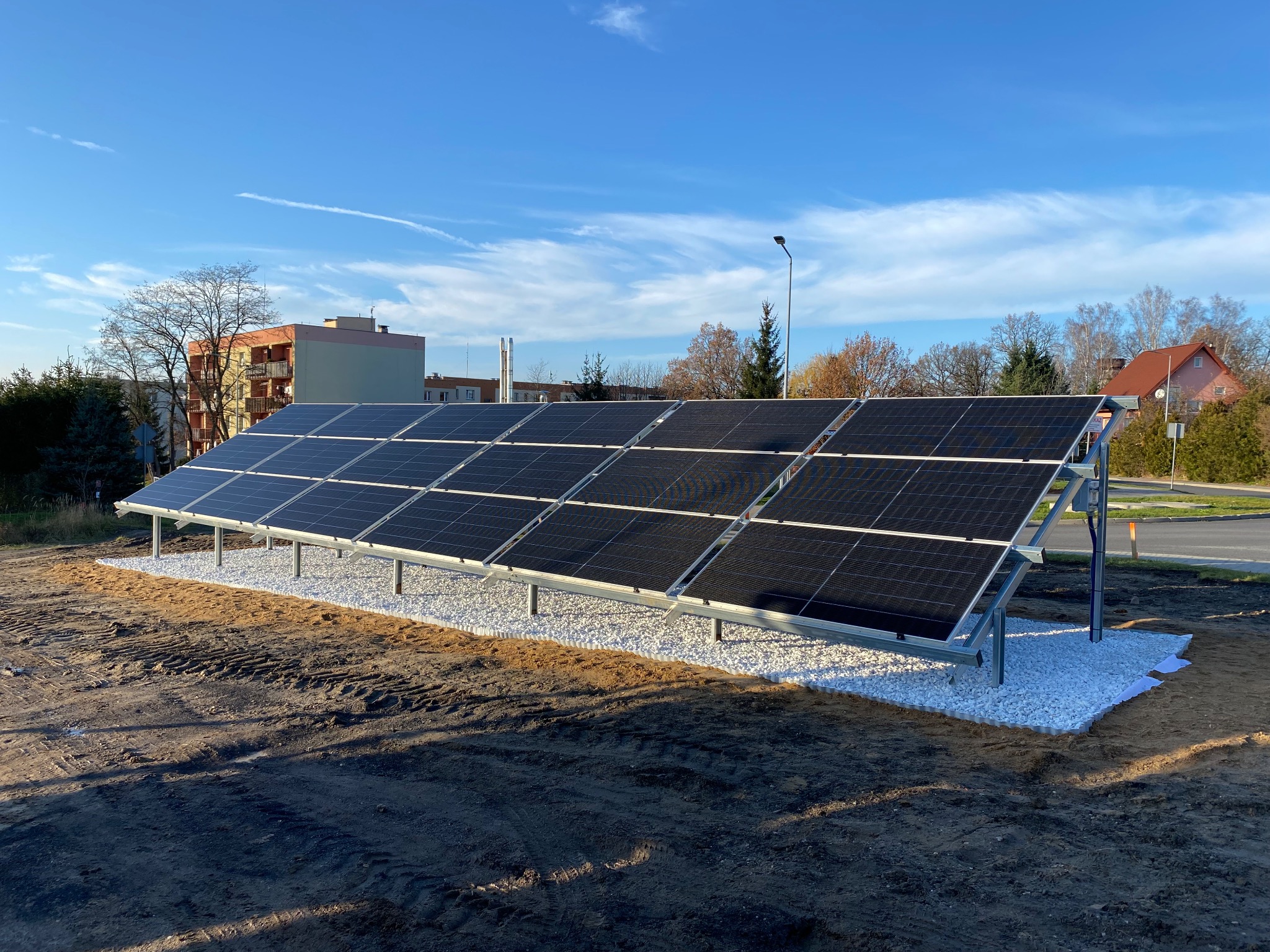 Rząd paneli słonecznych na metalowej konstrukcji, otoczony białym żwirem, na tle błękitnego nieba i zabudowań.
