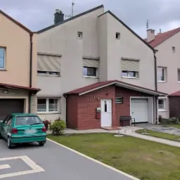 Szeregowy dom z jasną elewacją, czerwonym ceglanym gankiem i zielonym samochodem na podjeździe w pochmurny dzień.