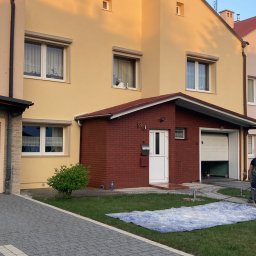 Mateusz Bubienczyk Usługi Budowlane - Dwukondygnacyjny dom w kolorze żółtym z czerwonym ceglanym garażem z numerem 13, widoczna plandeka na trawniku i fragment chodnika z kostki brukowej.