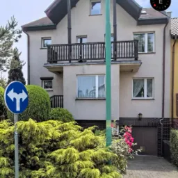 Dwupiętrowy dom z balkonem, widoczny z poziomu ulicy, z bujną zielenią w ogrodzie i znakiem drogowym na pierwszym planie.