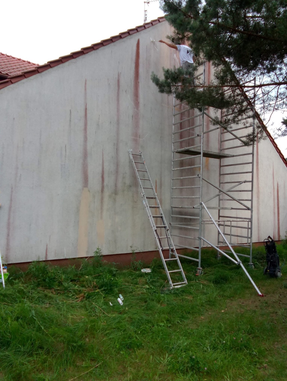 Pracownik na rusztowaniu maluje elewację budynku z widocznymi zaciekami, obok stoi drabina, na trawie leżą śmieci.