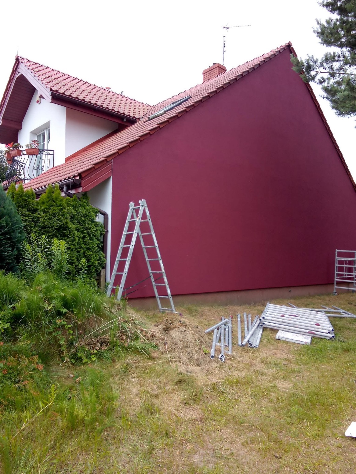 Ściana domu jednorodzinnego pomalowana na bordowo; widoczna drabina przystawiona do ściany, elementy rusztowania na trawie, fragment balkonu z kwiatami.