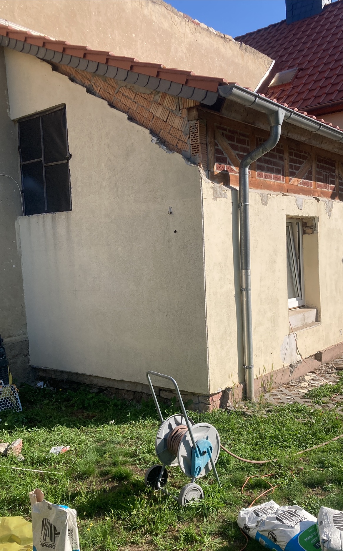 Fragment elewacji budynku w trakcie remontu, z widocznymi odsłoniętymi cegłami, częściowo otynkowaną ścianą, rynną, zwiniętym wężem na wózku i workami zaprawy na trawie.