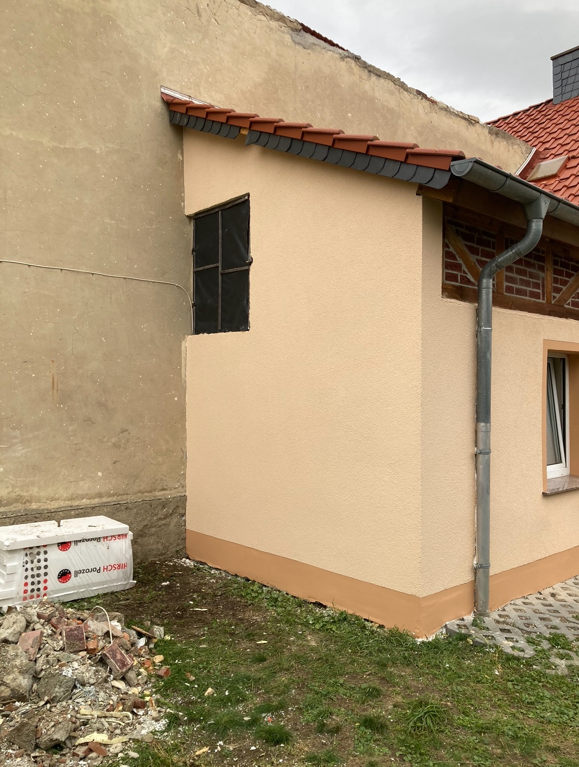 Świeżo otynkowana elewacja budynku z widocznym oknem zabezpieczonym folią, obok stary budynek z uszkodzonym tynkiem, na ziemi gruz i materiały budowlane.