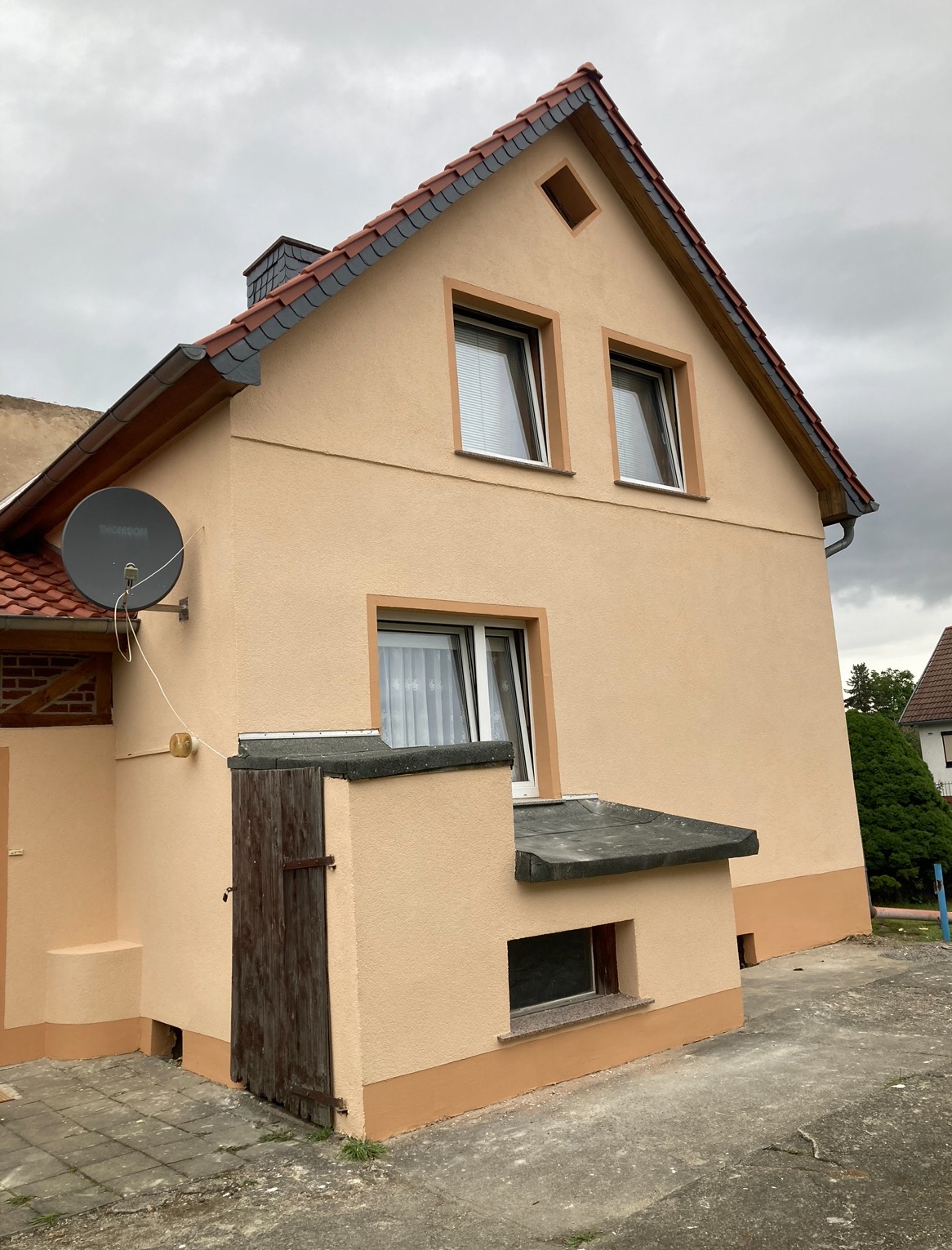 Dwukondygnacyjny dom z elewacją w odcieniach beżu, widoczny z boku, z anteną satelitarną na ścianie i drewnianymi drzwiami do piwnicy.