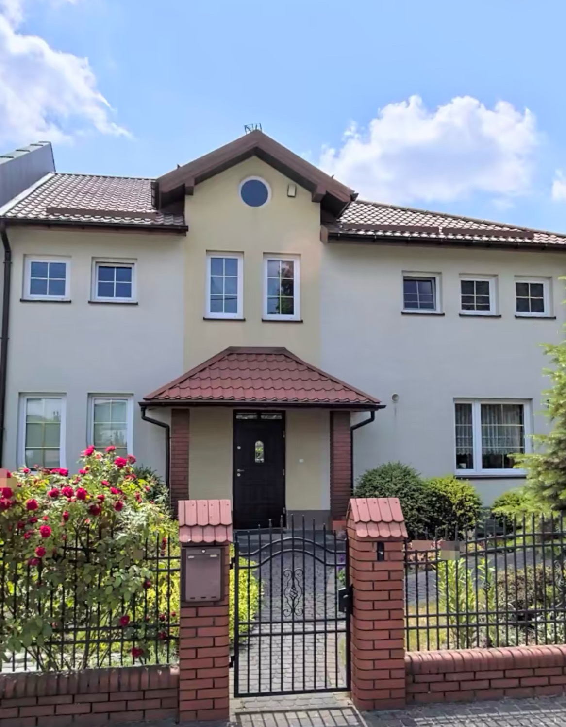 Dwupiętrowy dom z beżową elewacją, czerwoną dachówką i czarną bramą wjazdową, otoczony zielenią i kwitnącymi różami.