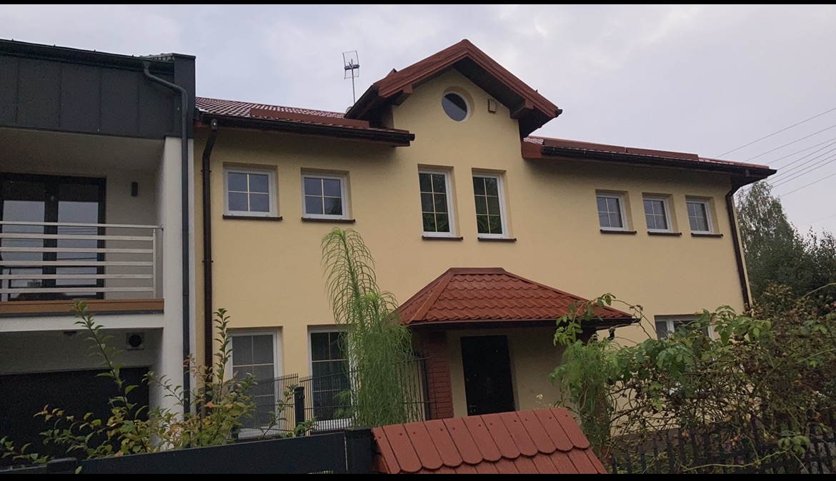 Dwukondygnacyjny dom z żółtą elewacją, brązowym dachem i anteną na szczycie, widoczny zza ogrodzenia porośniętego roślinnością.