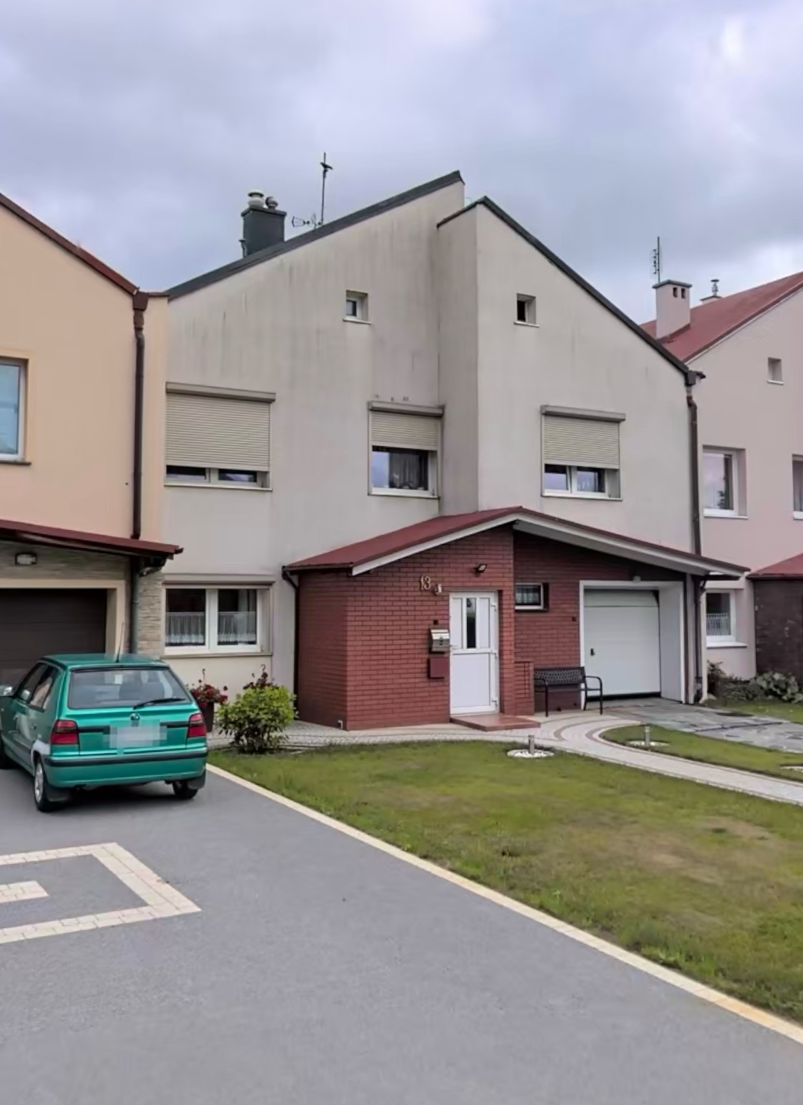 Szeregowy dom z jasną elewacją, czerwonym ceglanym gankiem i zielonym samochodem na podjeździe w pochmurny dzień.