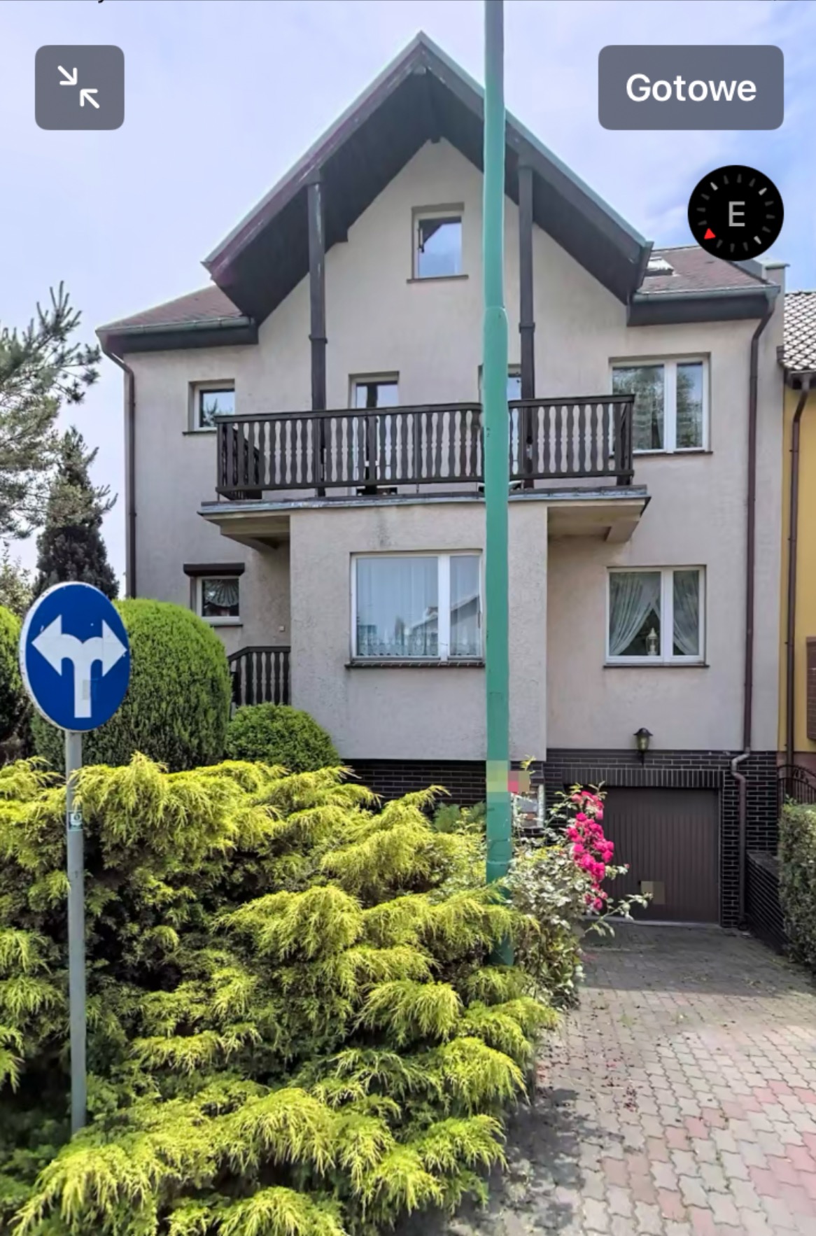 Dwupiętrowy dom z balkonem, widoczny z poziomu ulicy, z bujną zielenią w ogrodzie i znakiem drogowym na pierwszym planie.