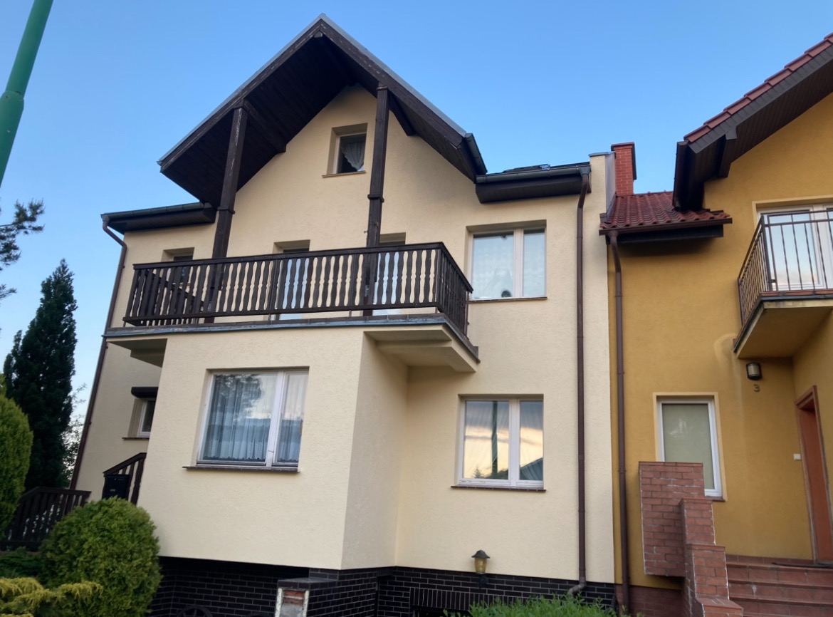 Jasna elewacja domu z ciemnobrązowymi elementami drewnianymi, balkon z drewnianą balustradą, widok z dołu na budynek sąsiadujący z żółtą elewacją.