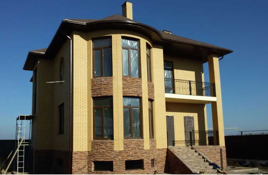 Dwupiętrowy dom z żółtej cegły z elementami dekoracyjnymi z kamienia, balkonem z kutą balustradą i oknami zabezpieczonymi folią ochronną, widziany w słoneczny dzień.