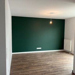 Yaroslav Danchenko. Usługi budowlane - Pomalowany na ciemnozielono pokój z drewnianą podłogą, białymi listwami przypodłogowymi i grzejnikiem, oświetlony pojedynczą żarówką na kablu.