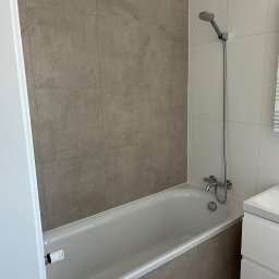 Yaroslav Danchenko. Usługi budowlane - Wykończona łazienka w Poznaniu: beżowe płytki imitujące kamień na ścianach i obudowie wanny, biała wanna z baterią i słuchawką prysznicową, biała szafka pod umywalką.