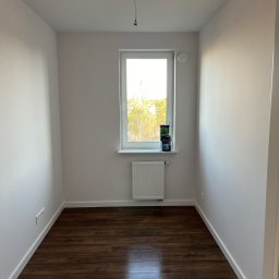 Yaroslav Danchenko. Usługi budowlane - Pusty pokój po remoncie z brązową podłogą z paneli, białymi ścianami i grzejnikiem pod oknem, na parapecie stoją puszki z farbą.