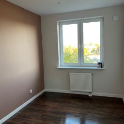 Yaroslav Danchenko. Usługi budowlane - Pokój po remoncie: ściana pomalowana na kolor terakoty, widoczne drewniane panele podłogowe, białe okno z widokiem na zieleń, biały grzejnik i puszka z farbą na parapecie.