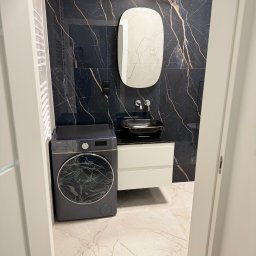 Yaroslav Danchenko. Usługi budowlane - Elegancka łazienka z czarnymi płytkami imitującymi marmur, owalnym lustrem i umywalką nablatową. Pralka w kolorze grafitowym. Całość w nowoczesnym stylu.