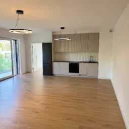 Nowoczesne wnętrze mieszkania z drewnianą podłogą, minimalistyczną kuchnią i czarną lodówką. Widok z balkonu w tle, oświetlenie punktowe.