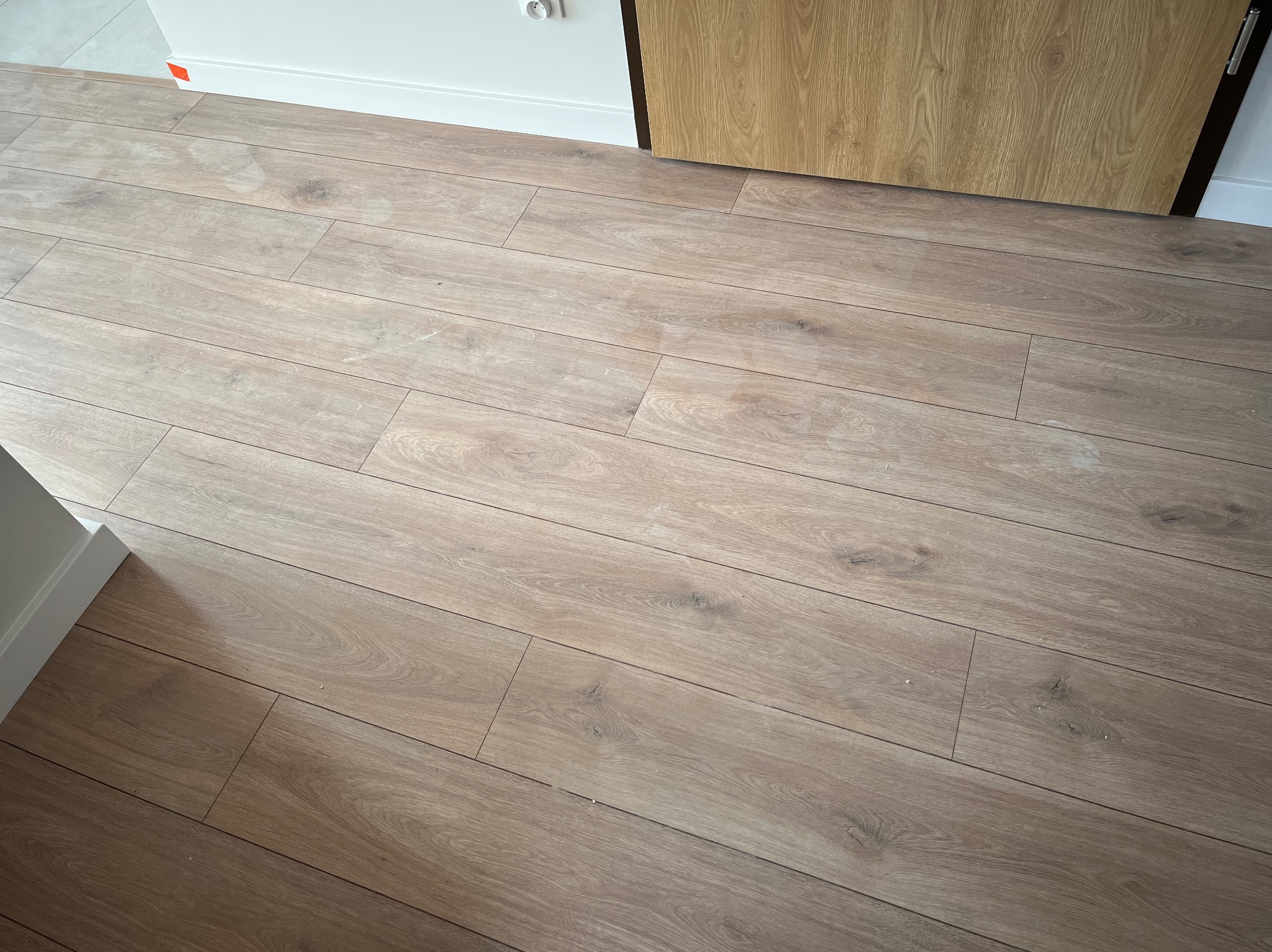 Jasne panele podłogowe ułożone wzdłuż ściany z białą listwą przypodłogową i fragmentem dębowych drzwi.