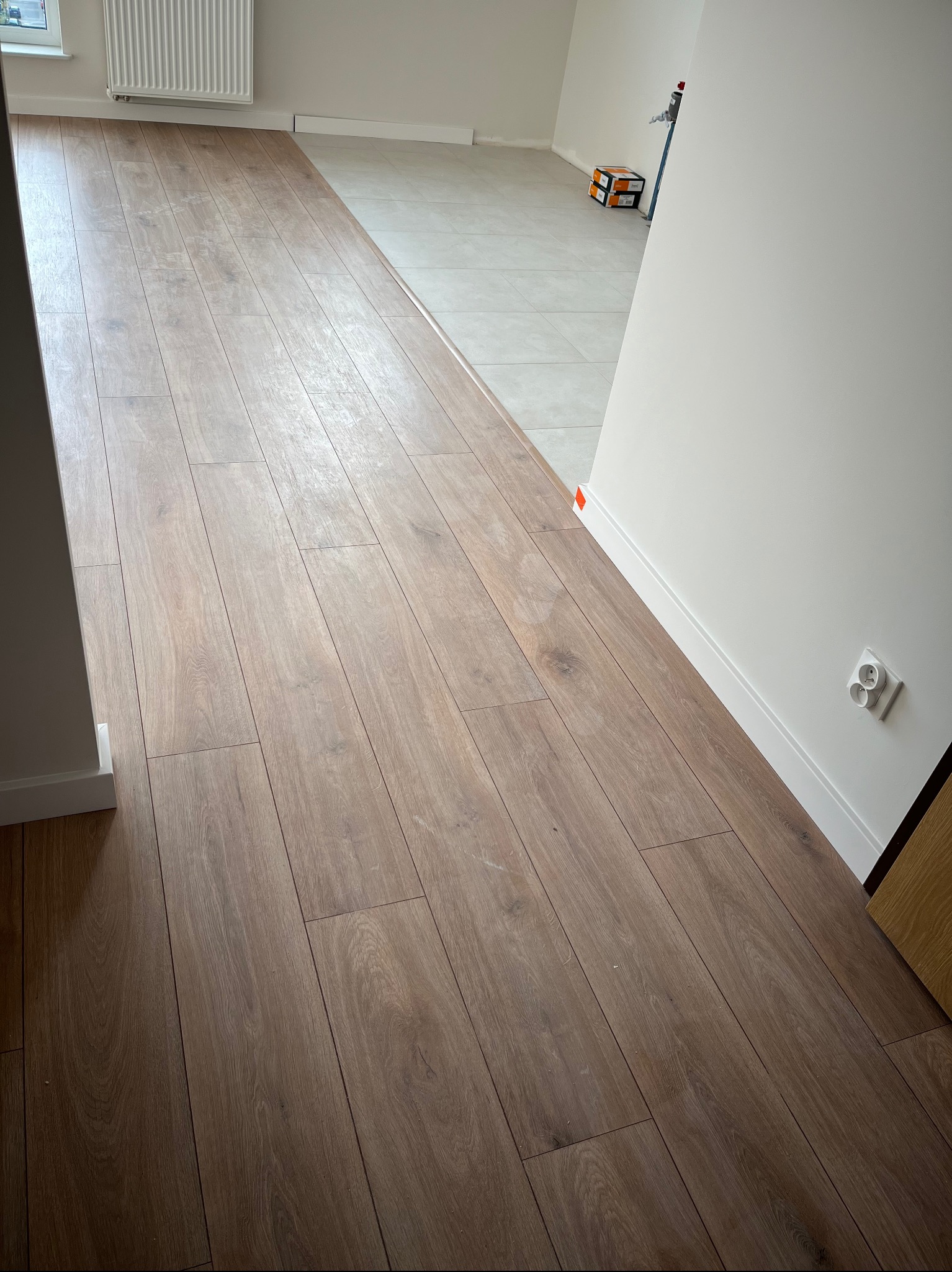 Fragment podłogi z paneli w odcieniach brązu, widoczny fragment płytek podłogowych w jasnym odcieniu, białe listwy przypodłogowe i gniazdko elektryczne na ścianie.