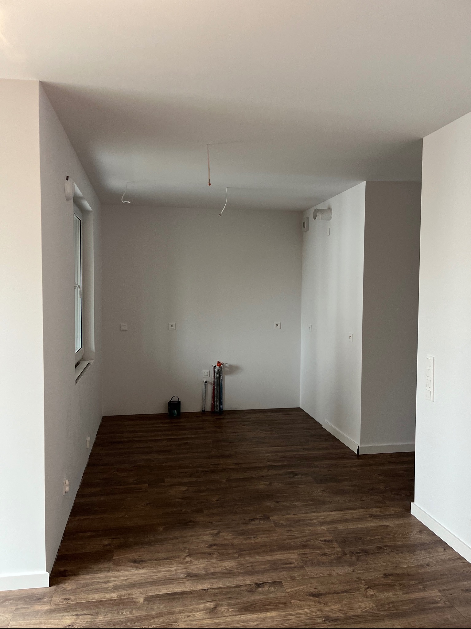 Pusty pokój po remoncie z ciemnymi panelami podłogowymi, widoczne wyprowadzenia instalacji elektrycznej i hydraulicznej, ściany pomalowane na biało.