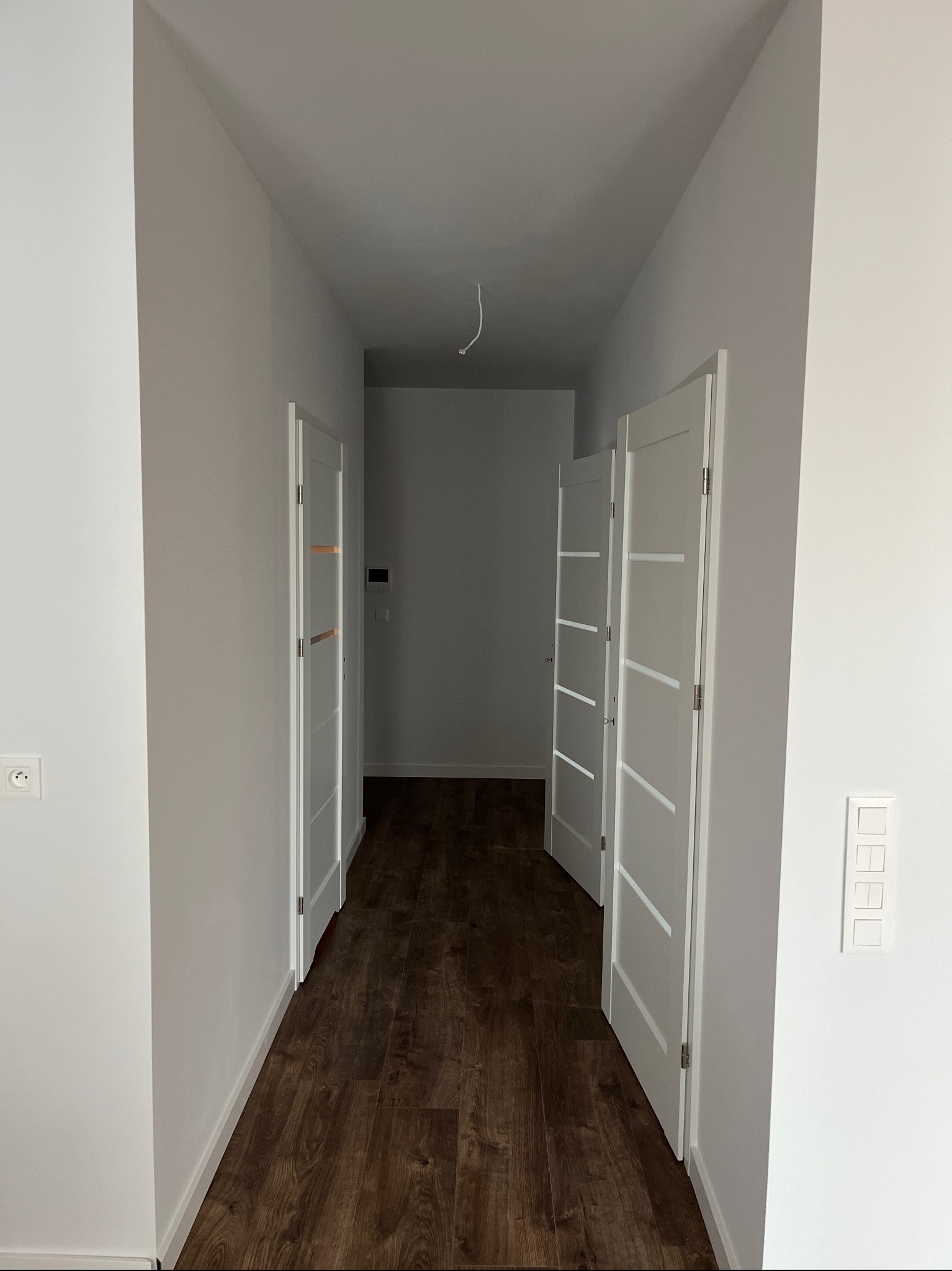 Korytarz w nowym mieszkaniu z dwiema parami białych drzwi z przeszkleniami i panelami podłogowymi w odcieniach brązu, widok z perspektywy wejścia.